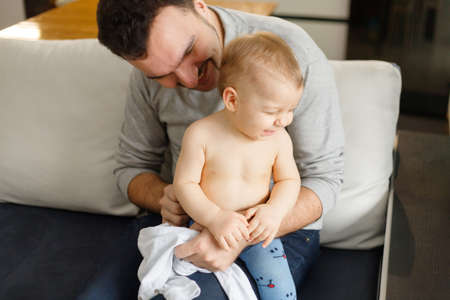 Father dresses infant in living room sitting on couch at home. Clothes baby undershirts and sliders on baby close-up. Home care and baby care concept.の写真素材