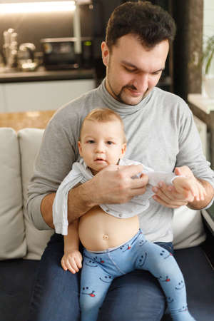 Father dresses infant in living room sitting on couch at home. Clothes baby undershirts and sliders on baby close-up. Home care and baby care concept.の写真素材