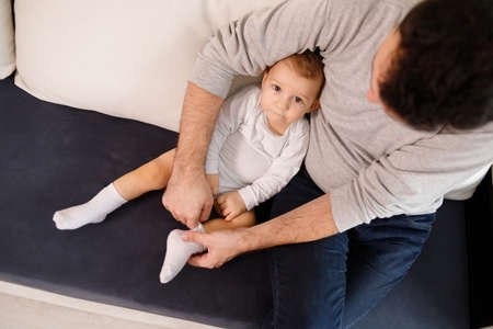Young father is putting socks on his baby son, sitting on sofaの写真素材
