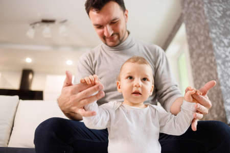 Young father assisting walking his infant son teaching to walk at home. Daddy helps son attempting to walkの写真素材
