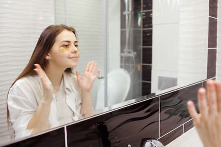 Caucasian woman applying hydrogel patches on under eye area, looking at mirror. Happy young lady enjoying morning anti age anti wrinkle routine in bathroomの写真素材