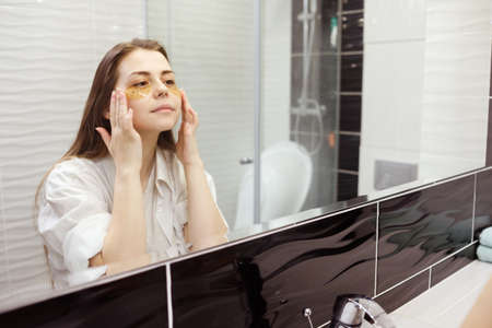 Caucasian woman applying hydrogel patches on under eye area, looking at mirror. Happy young lady enjoying morning anti age anti wrinkle routine in bathroom.の写真素材