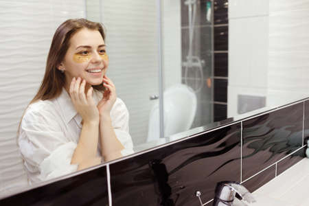 Caucasian woman applying hydrogel patches on under eye area, looking at mirror. Happy young lady enjoying morning anti age anti wrinkle routine in bathroomの写真素材