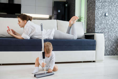 Baby boy play with mop, while mother surfing in your smartphone lying on couch.の写真素材