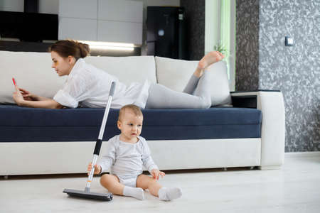 Baby boy play with mop, while mother surfing in your smartphone lying on couch.の写真素材