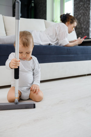 Baby boy play with mop, while mother surfing in your smartphone lying on couchの写真素材