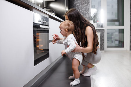 Happy young mum and cute kid daughter wear aprons bake muffins in kitchen, smiling mother teaching child take tray with cupcakes dessert make cakes out oven stove cook at home, family bakery conceptの写真素材