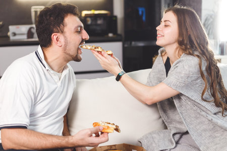 Happy couple at home feeding each other with pizza, dating at home. Care and love concept.の写真素材
