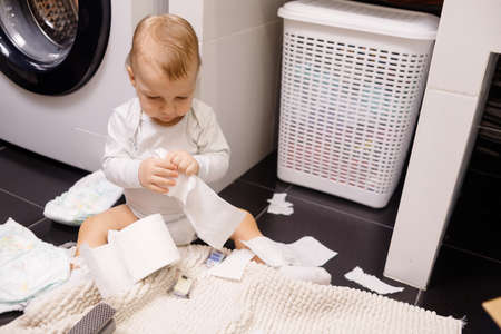the child sits on a white toilet. in his hands is toilet paper. there are a lot of rolls on the floor. close upの写真素材