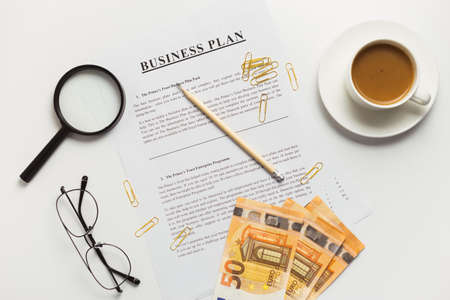 Flat lay of business plan documents with money, glasses, coffee cup, paper clip and supplies on white table desk.の写真素材