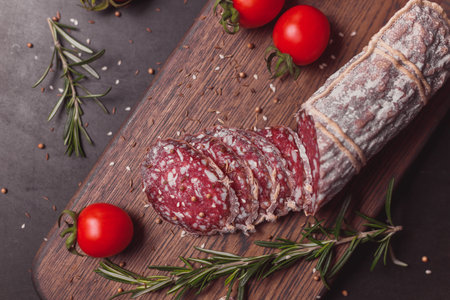 Smoked ham sausage and cutted slices with rosemary and cherry tomatoes on dark wooden boardの写真素材