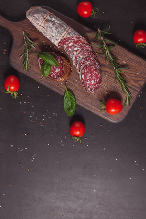 Smoked ham sausage and cutted slices with rosemary and cherry tomatoes on dark wooden board. Top view, flat layoutの写真素材
