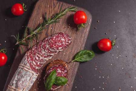 Smoked ham sausage and cutted slices with rosemary and cherry tomatoes on dark wooden boardの写真素材