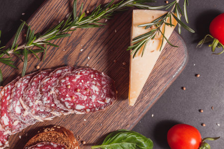 Smoked ham sausage and cutted slices with rosemary and cherry tomatoes on dark wooden board. Top view, flat layoutの写真素材