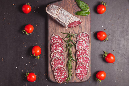 Smoked ham sausage and cutted slices with rosemary and cherry tomatoes on dark wooden board. Top...