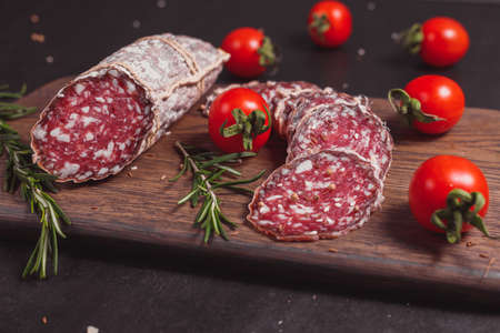 Smoked ham sausage and cutted slices with rosemary and cherry tomatoes on dark wooden boardの写真素材