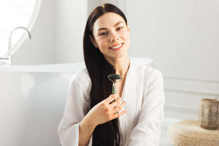 Beautiful girl holding drainage massage face roller sitting in luxury bathroom.の写真素材