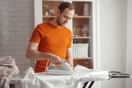 Young caucasian man ironing children's sheet on ironing board at home. Men doing home choresの写真素材