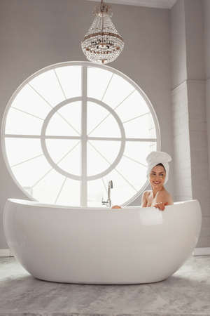Young woman relaxing lying in bathtube in luxury bathroom with round large window.の写真素材