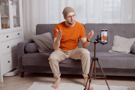 Millennial hipster man blogger recording vlog on mobile phone sit on sofa in living room and explain and gesturing with hands. Doing content for social media.の写真素材