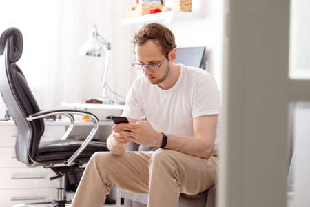 Happy young man in eyeglasses sitting on sofa sending text on mobile phoneの写真素材
