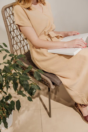 Woman reading book in wicker chair with home plant next to it. Girl wears beige dressの写真素材