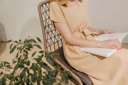 Woman reading book in wicker chair with home plant next to it. Girl wears beige dressの写真素材