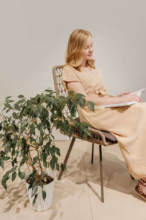 Woman reading book in wicker chair with home plant next to it. Girl wears beige dressの写真素材