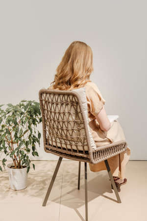 Woman reading book in wicker chair with home plant next to it. Girl wears beige dress. Back viewの写真素材