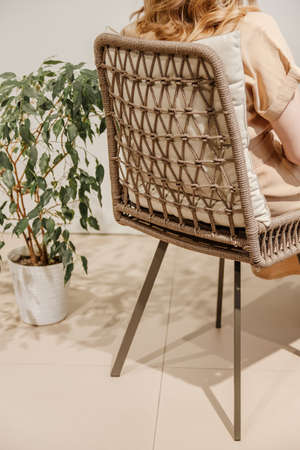 Woman reading book in wicker chair with home plant next to it. Girl wears beige dress. Back viewの写真素材