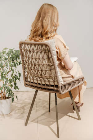 Woman reading book in wicker chair with home plant next to it. Girl wears beige dress. Back viewの写真素材