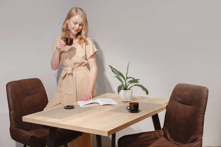 Minimalist interior. Woman in dress standing by table, drink tea and leaves through a bookの写真素材