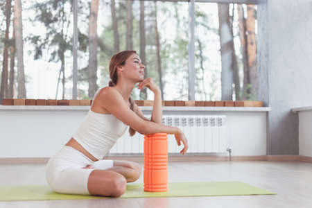 Sporty woman with foam roller sitting on mat in sport hall.の写真素材