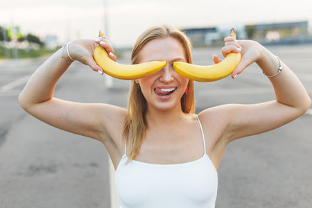 Frivolously and mischievous young woman with braces on teeth holding bananas on her face has fun outdoors. Emotionsの写真素材