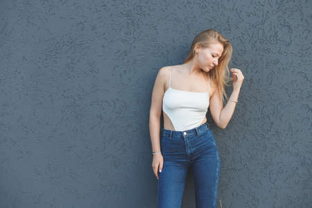 young blonde woman in the white sleeveless t-shirt and blue jeans standing over gray wall backgroundの写真素材