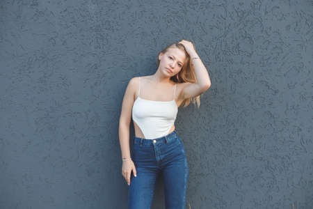 young blonde woman in the white sleeveless t-shirt and blue jeans standing over gray wall backgroundの写真素材