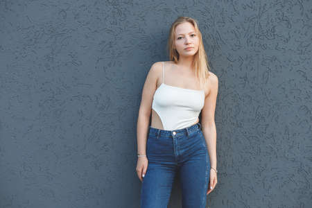 young blonde woman in the white sleeveless t-shirt and blue jeans standing over gray wall backgroundの写真素材