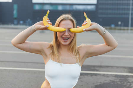Frivolously and mischievous young woman with braces on teeth holding bananas on her face has fun outdoorsの写真素材