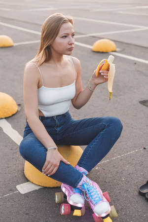 Beautiful cute bright girl on a turquoise background, laughs, holds a bunch of bananas and eats them, fashionable funny look, dressed in overalls in bananas, perfect make-upの写真素材