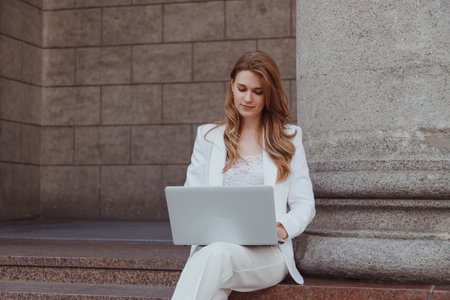Busy business woman in white suit with laptop sitting outdoors at classical pillar. Investing, working remotelyの写真素材