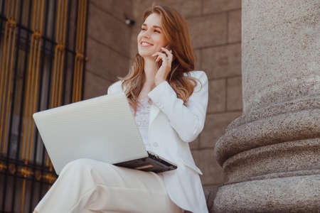 Long haired smiling cheerful lady in white t-shirt and jeans suit speaking on mobile phone in outdoor terrace in street cafeの写真素材