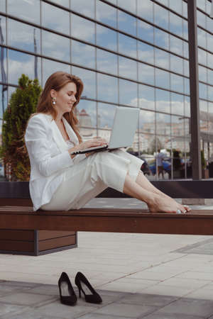Barefoot businesswoman in white suit sitting on bench with laptop.の写真素材