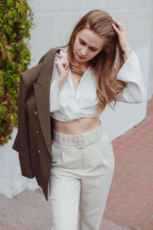 Fashionable woman in white pants and blouse with coat on shoulder posing outdoors. women autumn lookの写真素材