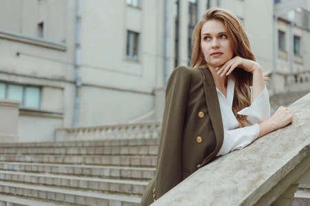 Fashionable woman in white pants and blouse with coat on shoulder posing outdoors. women autumn lookの写真素材