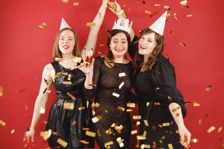 Three delighted women in black dress and party hats over red background with golden confetti. female partyの写真素材