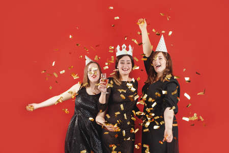 Three delighted women in black dress and party hats over red background with golden confetti. female partyの写真素材