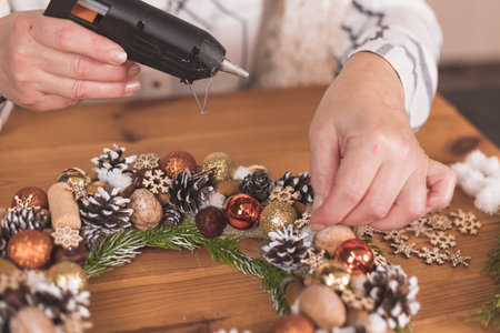 Close-up of female hands glues toys to a Christmas wreath on a wooden table. concept for New Year,...