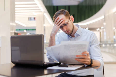 Young puzzled businessman working in empty food court in mall with documents and laptop. Filing tax returns work with contractsの写真素材