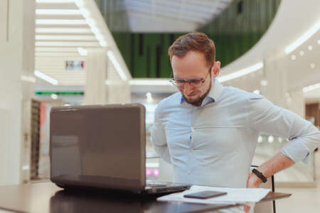 Man holds on to his back, suffers from lower back pain, working on computer. Sedentary work office workerの写真素材