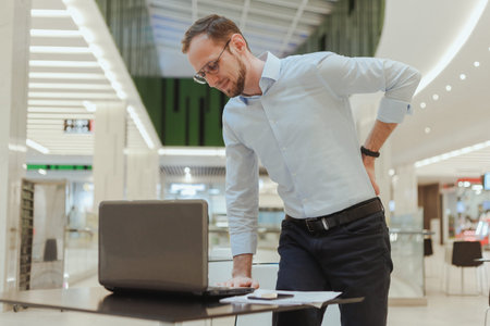 A man holds on to his back, suffers from lower back pain, standing near a desktop with a computer....